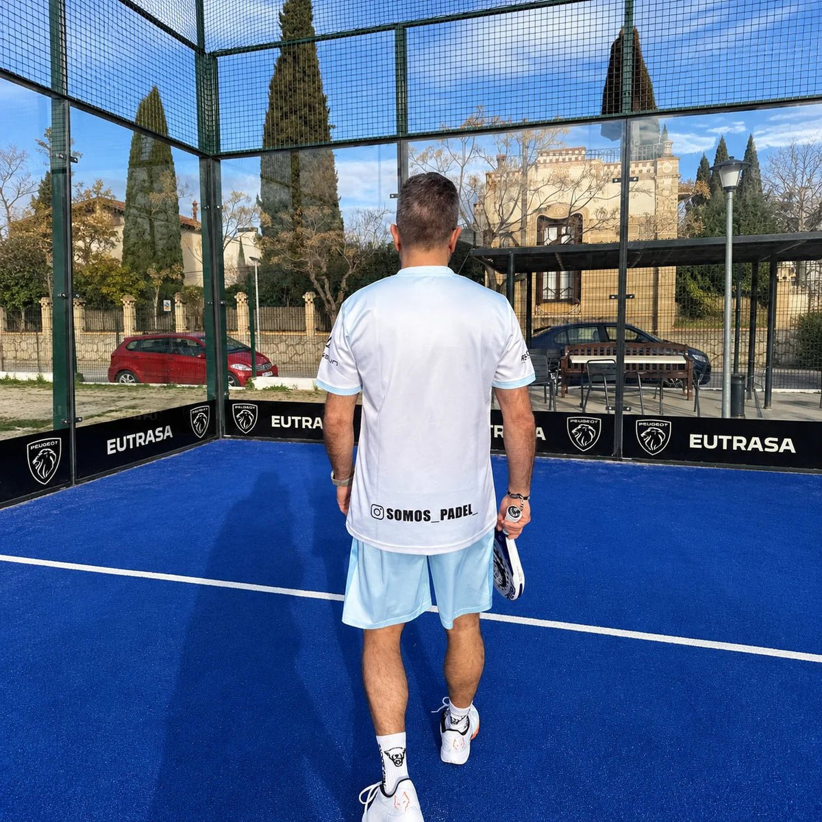 Vista trasera de la equipación de pádel blanca y celeste con logotipo del club en la espalda