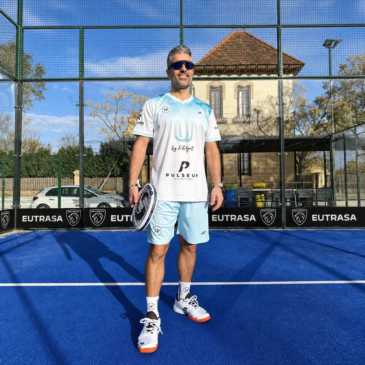 Jugador con equipación de pádel blanca y celeste personalizada y pala, de pie en pista azul