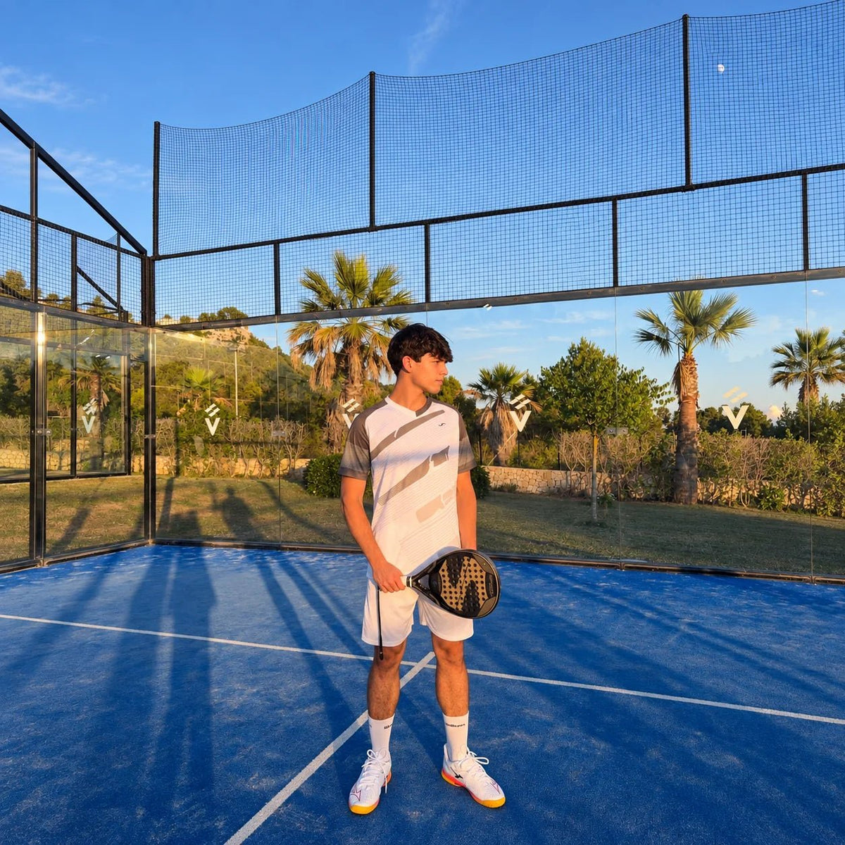 Jugador con conjunto de pádel blanco y gris y pala, de cuerpo entero en pista azul