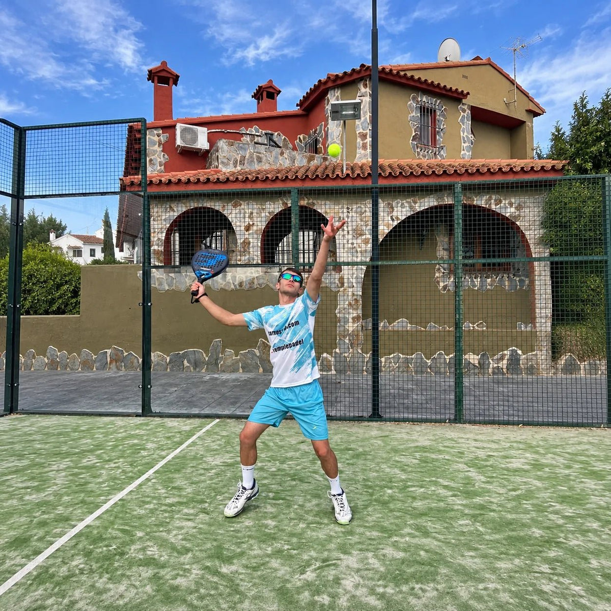 Jugador con equipación de pádel celeste saltando para golpear la bola en pista exterior