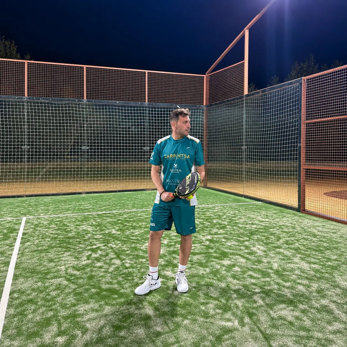 Jugador con equipación de pádel verde petróleo en posición de espera en pista de césped